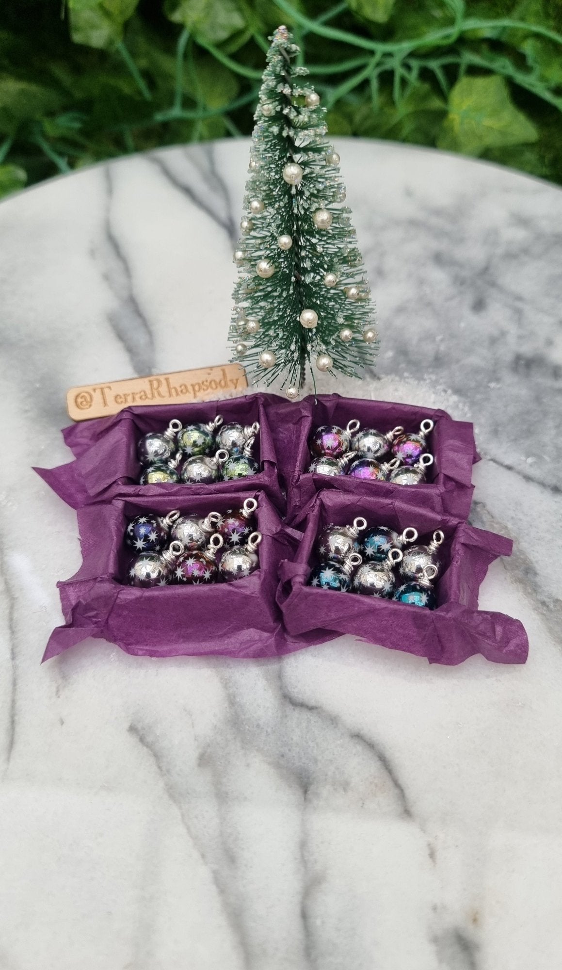 4 miniature 1:12 scale open tray boxes contain 6 Christmas baubles. The baubles are multiple colours with star motifs. The boxes are lined with purple paper displayed in front of a Christmas tree. @TerraRhapsody is written on wood in the background