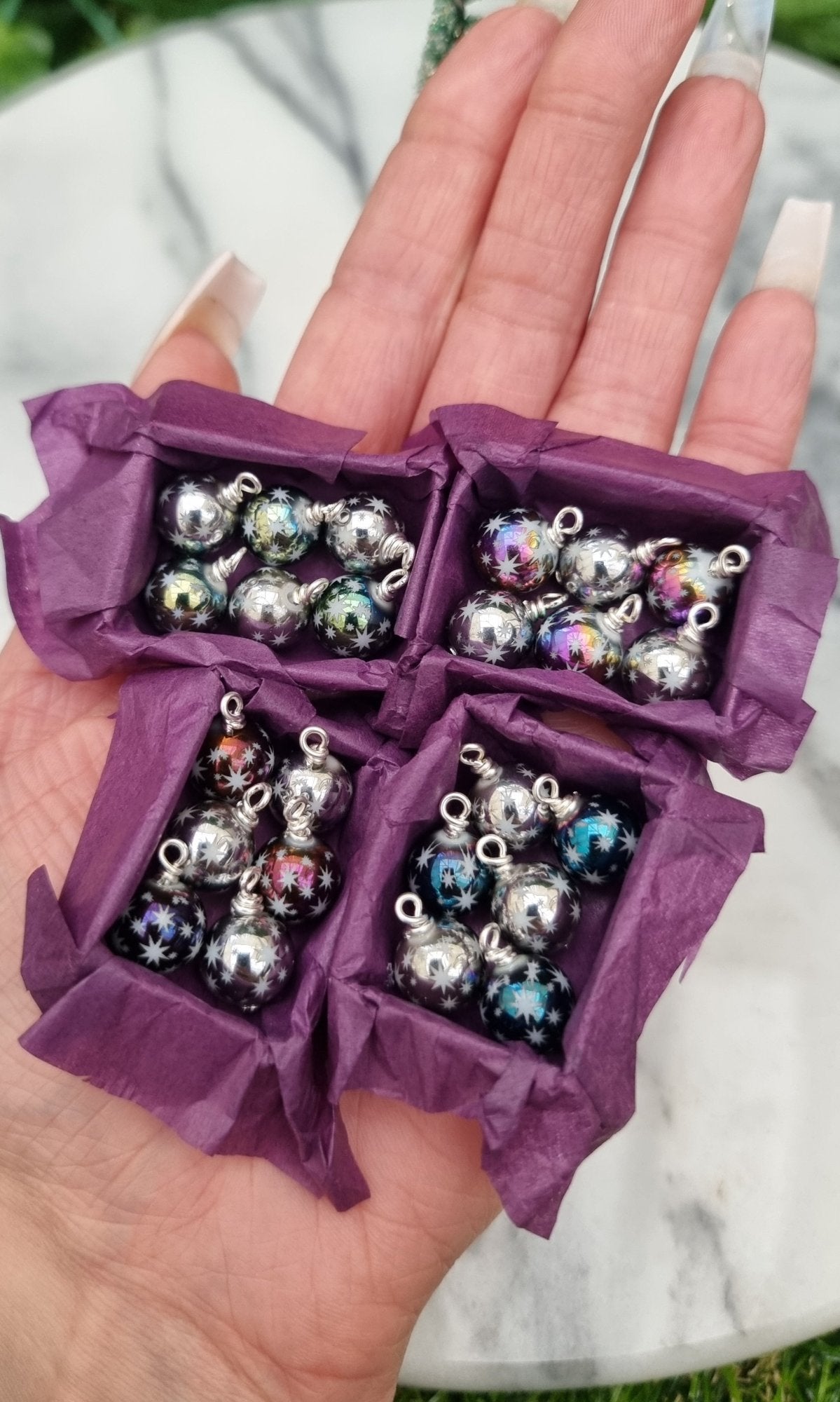 4 miniature 1:12 scale open tray boxes contain 6 Christmas baubles. The baubles are multiple colours with star motifs. The boxes are lined with purple paper displayed on the palm of a hand