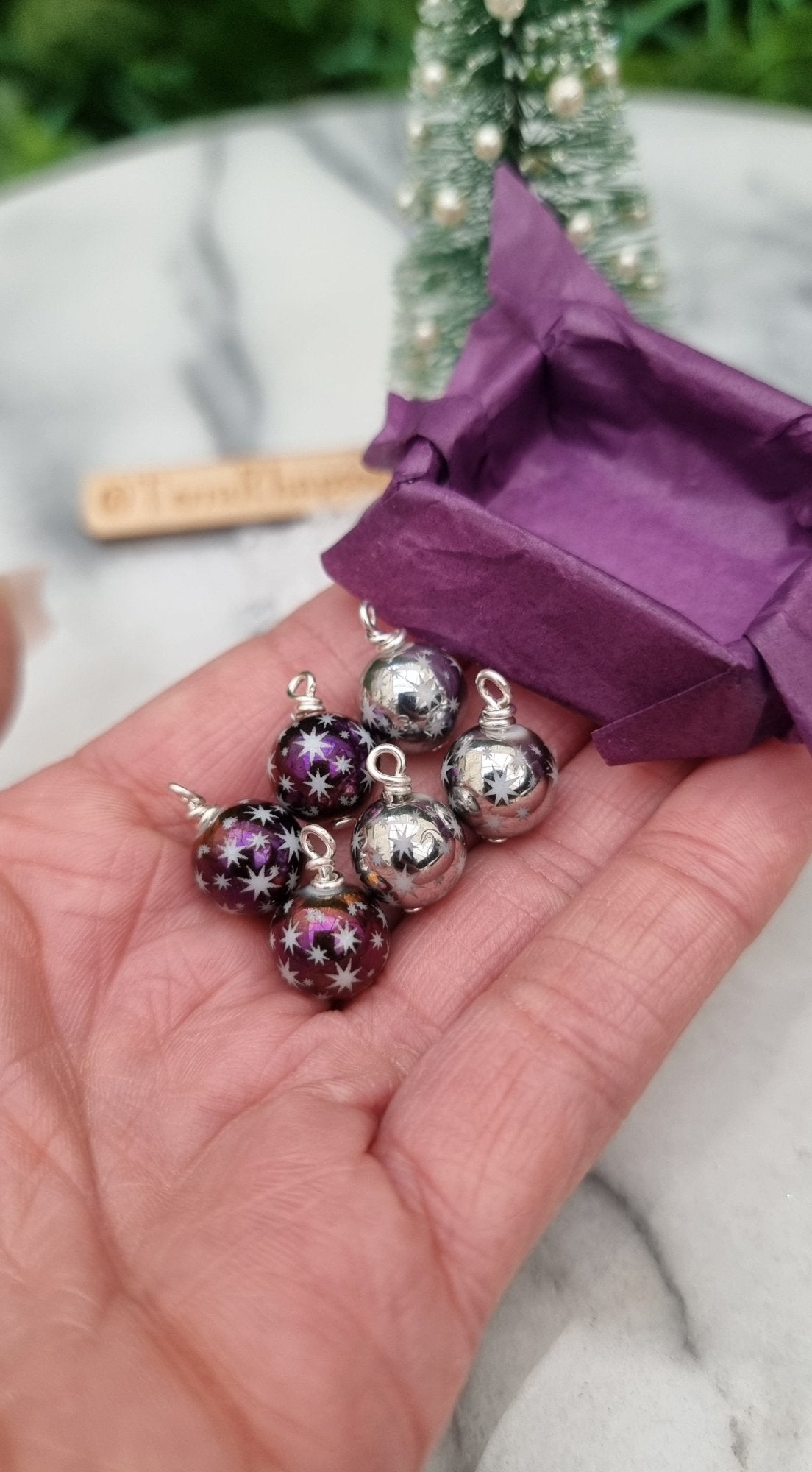 6 Miniature 1:12 Christmas baubles are displayed in an open hand with a purple lined tray. The baubles are silver and metallic purple coloured with white star motifs. @TerraRhapsody is written on wood in the background