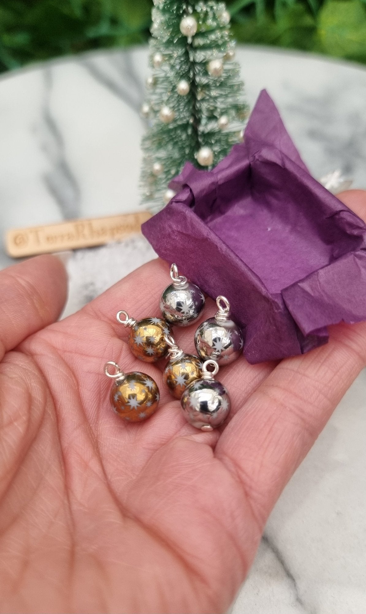 6 Miniature 1:12 Christmas baubles are displayed in an open hand with a purple lined tray. The baubles are silver and gold coloured with white star motifs. @TerraRhapsody is written on wood in the background