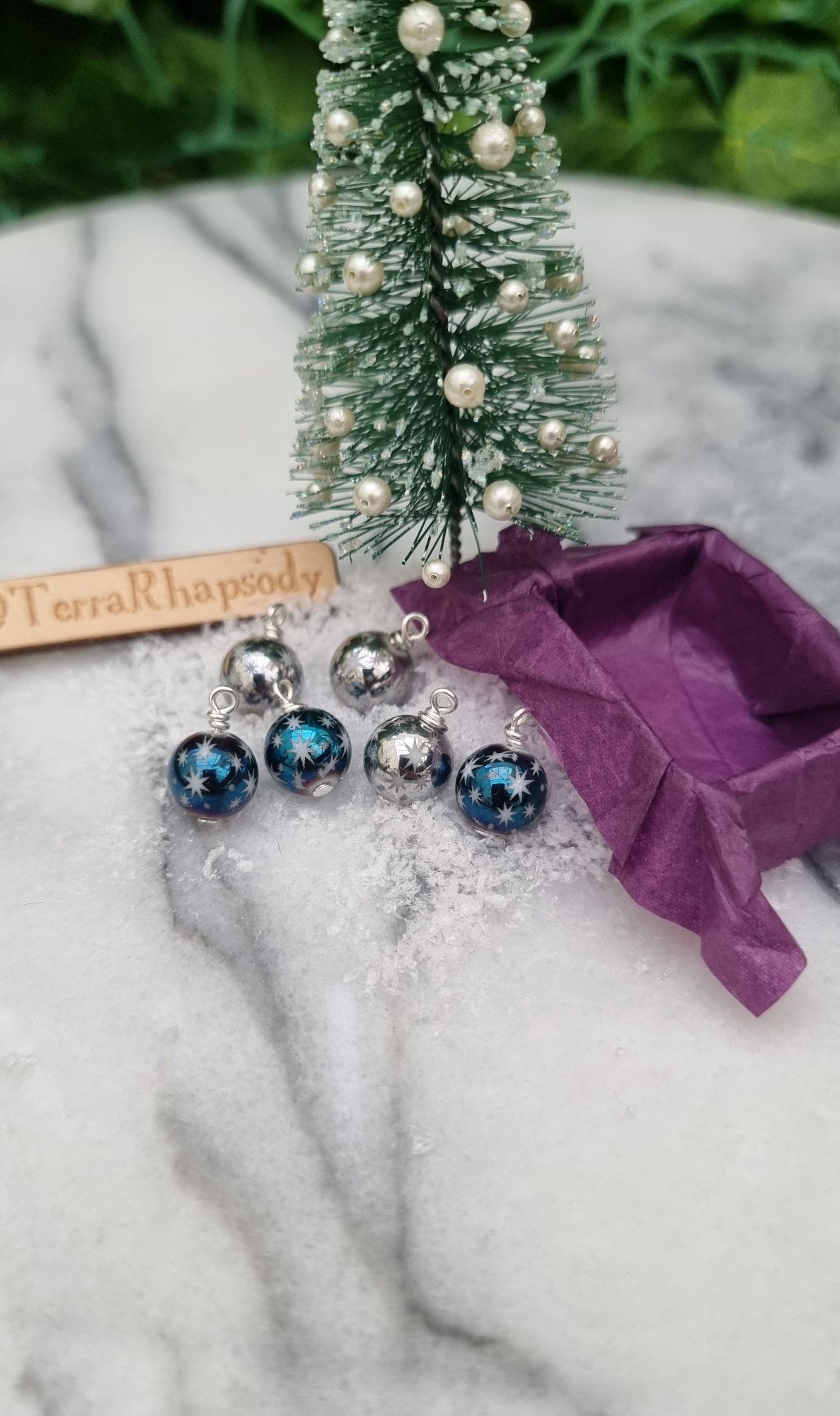 6 Miniature 1:12 Christmas baubles are displayed on snow beside a purple lined tray in front of a Christmas tree. The baubles are silver and metallic blue coloured with white star motifs. @TerraRhapsody is written on wood in the background