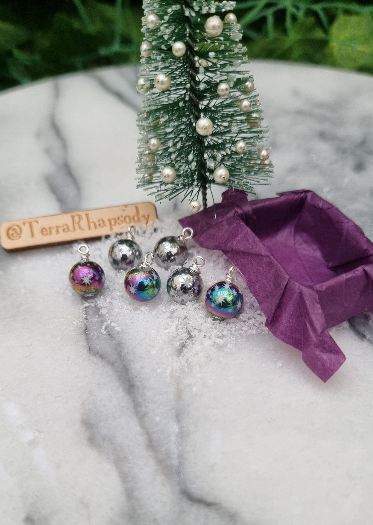 6 Miniature 1:12 Christmas baubles are displayed on snow beside a purple lined tray in front of a Christmas tree. The baubles are silver and rainbow coloured with white star motifs. @TerraRhapsody is written on wood in the background