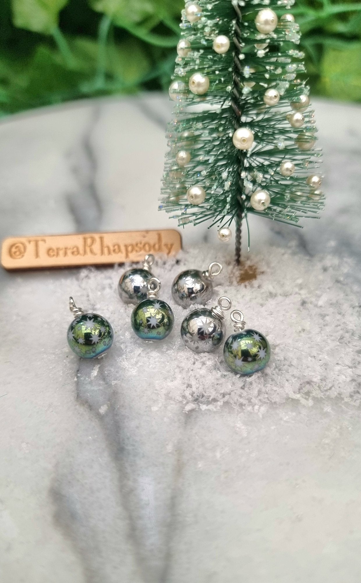 6 Miniature 1:12 Christmas baubles are displayed on snow tray in front of a miniature Christmas tree with pearl baubles. The baubles are silver and metallic pastel green coloured with white star motifs. @TerraRhapsody is written on wood in the background