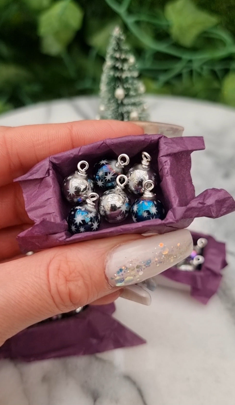 Miniature 1:12 scale open tray box contains 6 Christmas baubles. The baubles are silver and metallic blue coloured with white star motifs. The box is lined with purple paper, displayed between two fingers in front of a Christmas tree