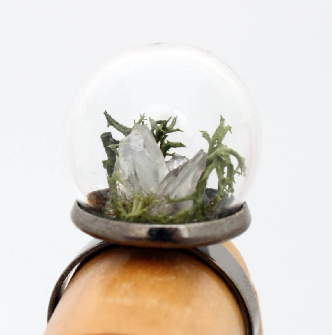 A wooden jointed hand displays this glass globe ring on a gunmetal ring base containing a crystal of lemurian seed quartz. The quartz is accompanied by reindeer moss and greenery inside the dome and displayed against a white background