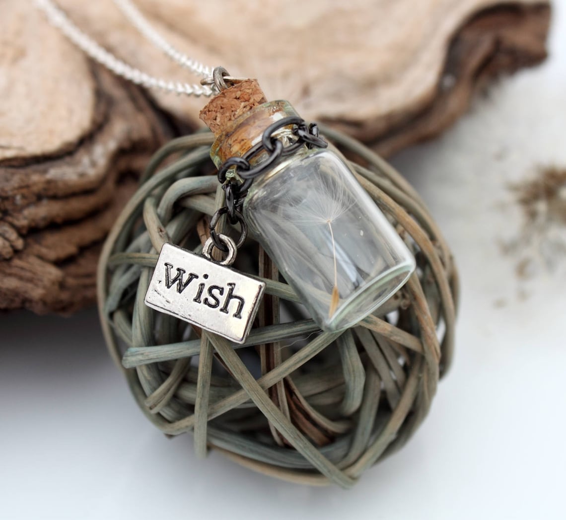 Miniature jar shaped glass vial necklace containing a dandelion seed with a silver coloured wish charm hung on black chain around the neck of the jar. Displayed a silver coloured chain attached to a cork lid