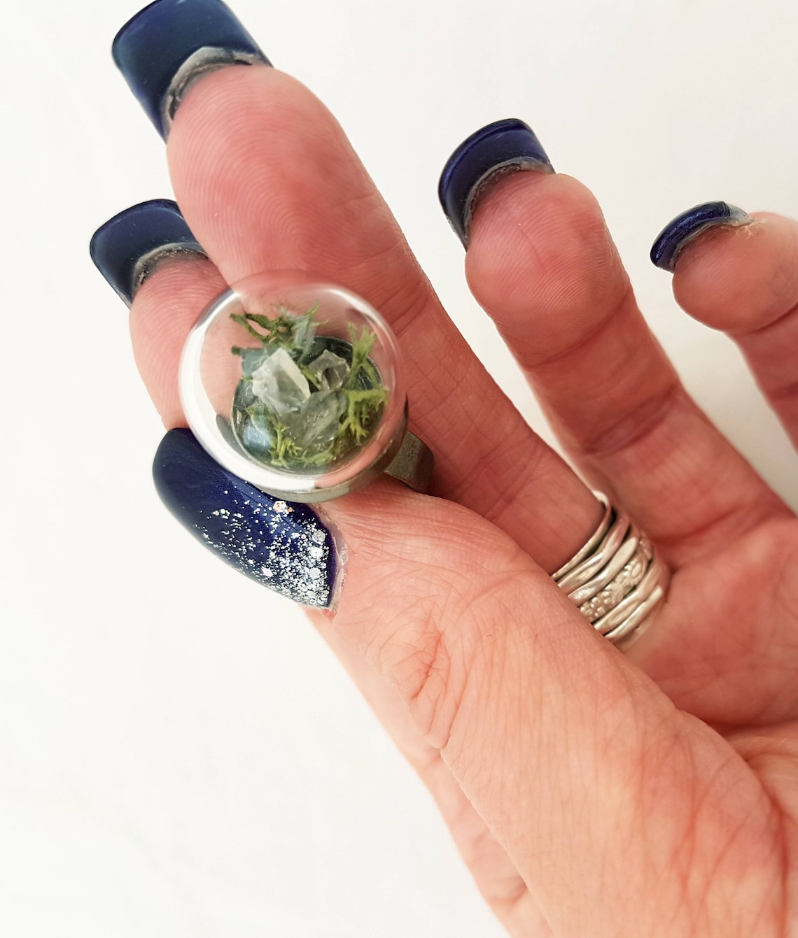 A hand displays this glass globe ring on a gunmetal ring base containing a crystal of lemurian seed quartz. The quartz is accompanied by reindeer moss and greenery inside the dome and displayed against a white background