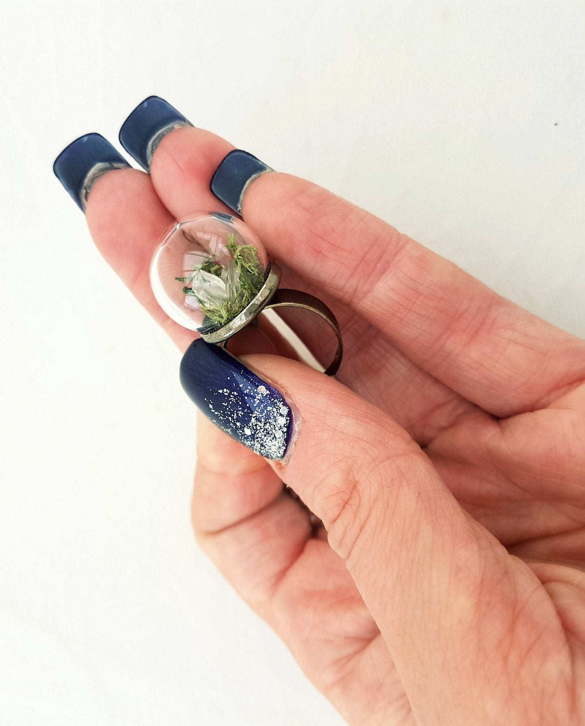 A hand displays this glass globe ring on a gunmetal ring base containing a crystal of lemurian seed quartz. The quartz is accompanied by reindeer moss and greenery inside the dome and displayed against a green background