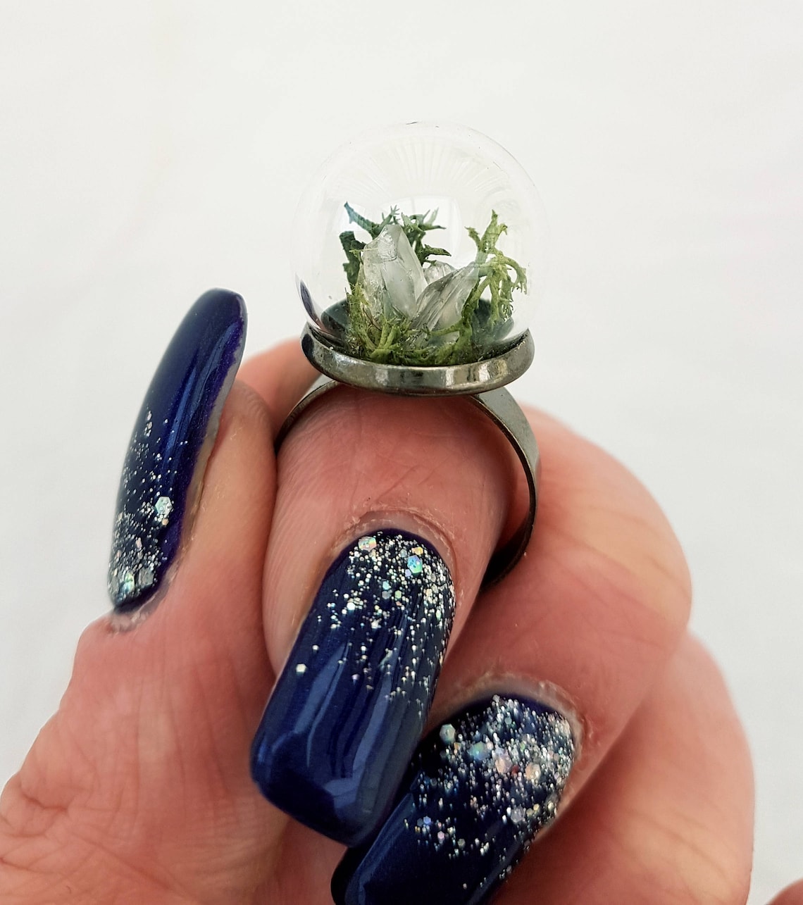 A hand displays this glass globe ring on a gunmetal ring base containing a crystal of lemurian seed quartz. The quartz is accompanied by reindeer moss and greenery inside the dome and displayed against a white background