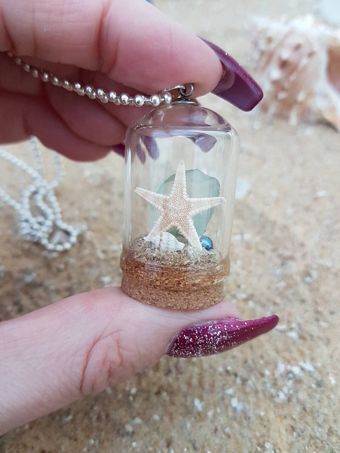 Hand holds a small glass bell jar with cork base and necklace attachment containing a starfish, shell, pearl, light green seaglass and greenery. It sits on a piece of aquamarine seaglass and sand