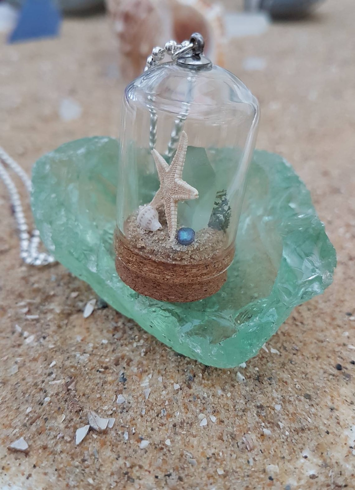 Small glass bell jar with cork base and necklace attachment containing a starfish, shell, pearl, light green seaglass and greenery. It sits on a piece of aquamarine seaglass and sand