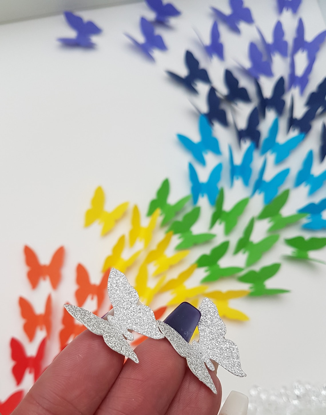 Close up of two silver glitter butterflies held on finger tips with Square white box frame showing a flurry of paper butterflies in rainbow colours, moving from red butterflies in the bottom left of the frame, through rainbow colours to the top.