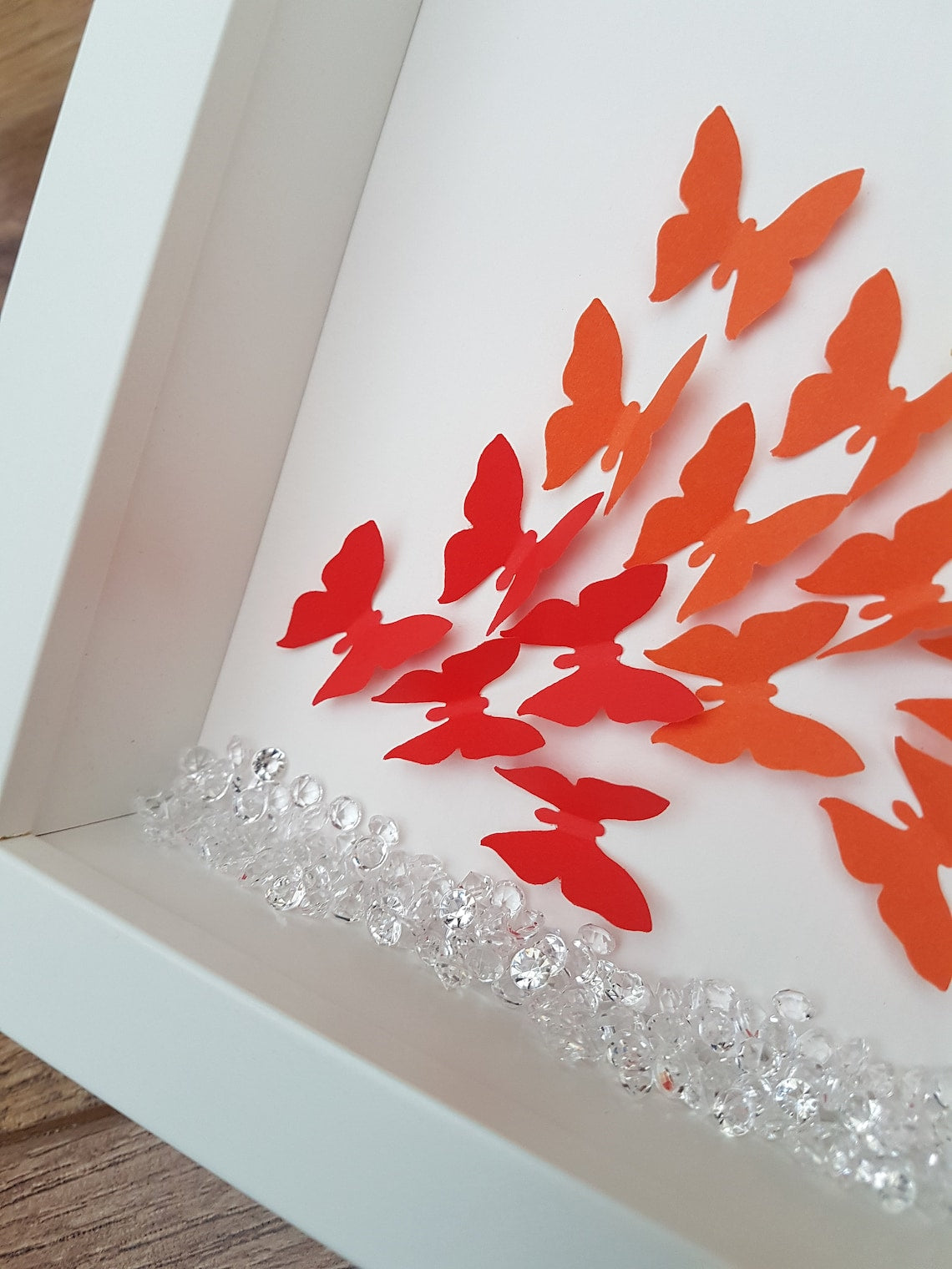 Square white box frame showing detail of a flurry of paper butterflies in rainbow colours, showing red  and orange butterflies in the bottom left of the frame, with plastic scatter crystals at the base of the frame