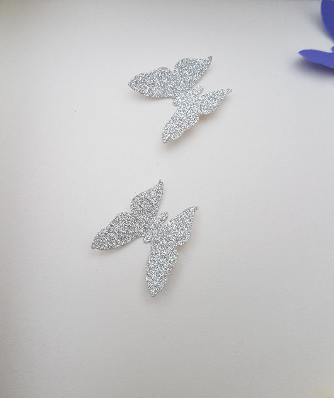 Two silver glitter coloured paper butterflies are shown on a white background