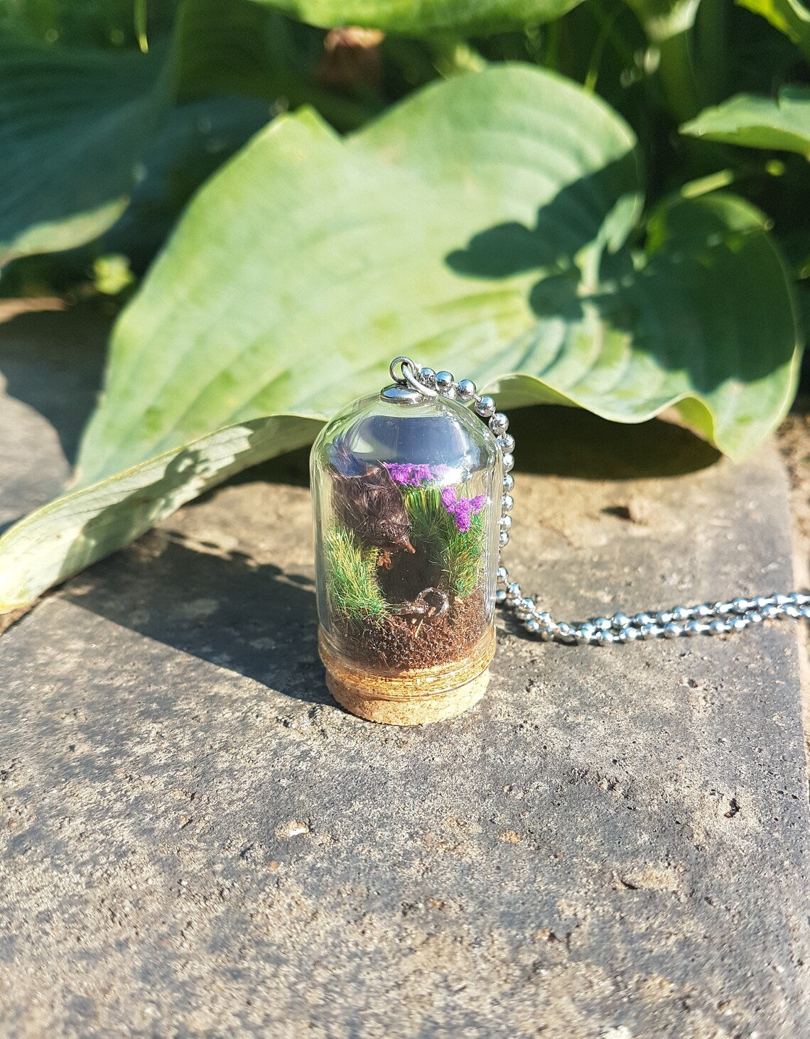 Miniature diorama glass bell jar necklace with cork base contains a tiny brown Robin. The Robin is surrounded by grass and purple flowers looking into an earthy hole containing a rusty key. The necklace is displayed on a grey stone with green leaves