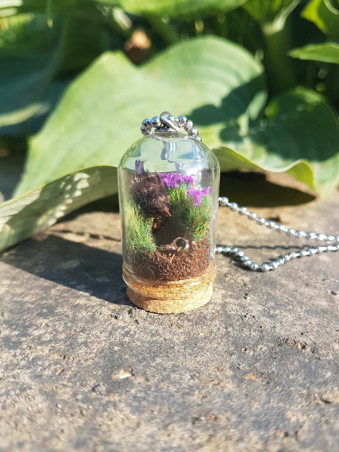 Miniature diorama glass bell jar necklace with cork base contains a tiny brown Robin. The Robin is surrounded by grass and purple flowers looking into an earthy hole containing a rusty key. The necklace is displayed on a grey stone with green leaves