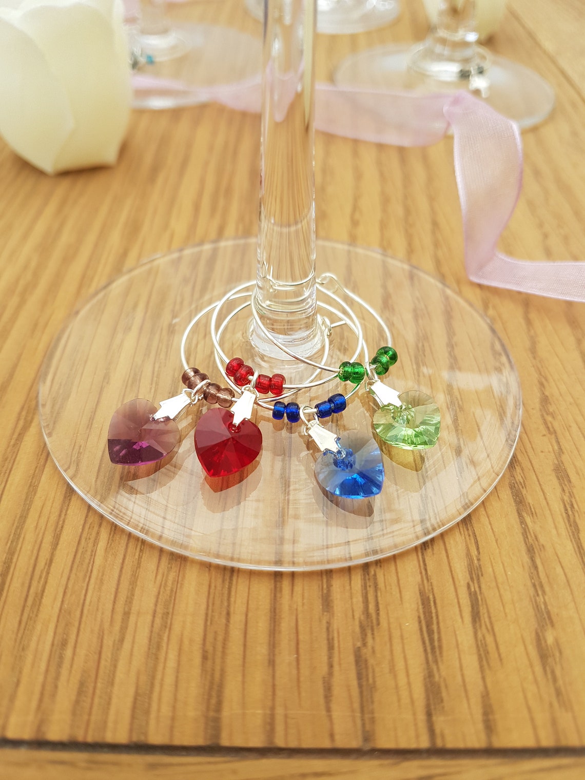 3 wine glass charms are arranged on top of  wine glass bases on a wooden background. The wine glass charms are round wire hoops, each with different coloured seed beads and a Swarovski crystal heart in red, green and lavender