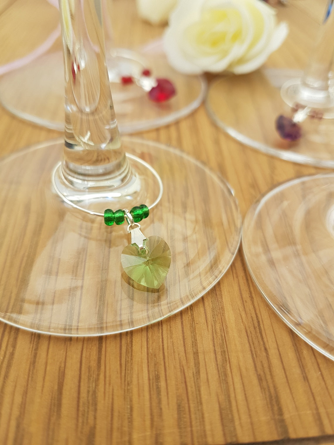 3 wine glass charms are arranged on top of wine glass bases on a wooden background. The wine glass charms are round wire hoops, each with different coloured seed beads and a Swarovski crystal heart in red, green, blue and lavender