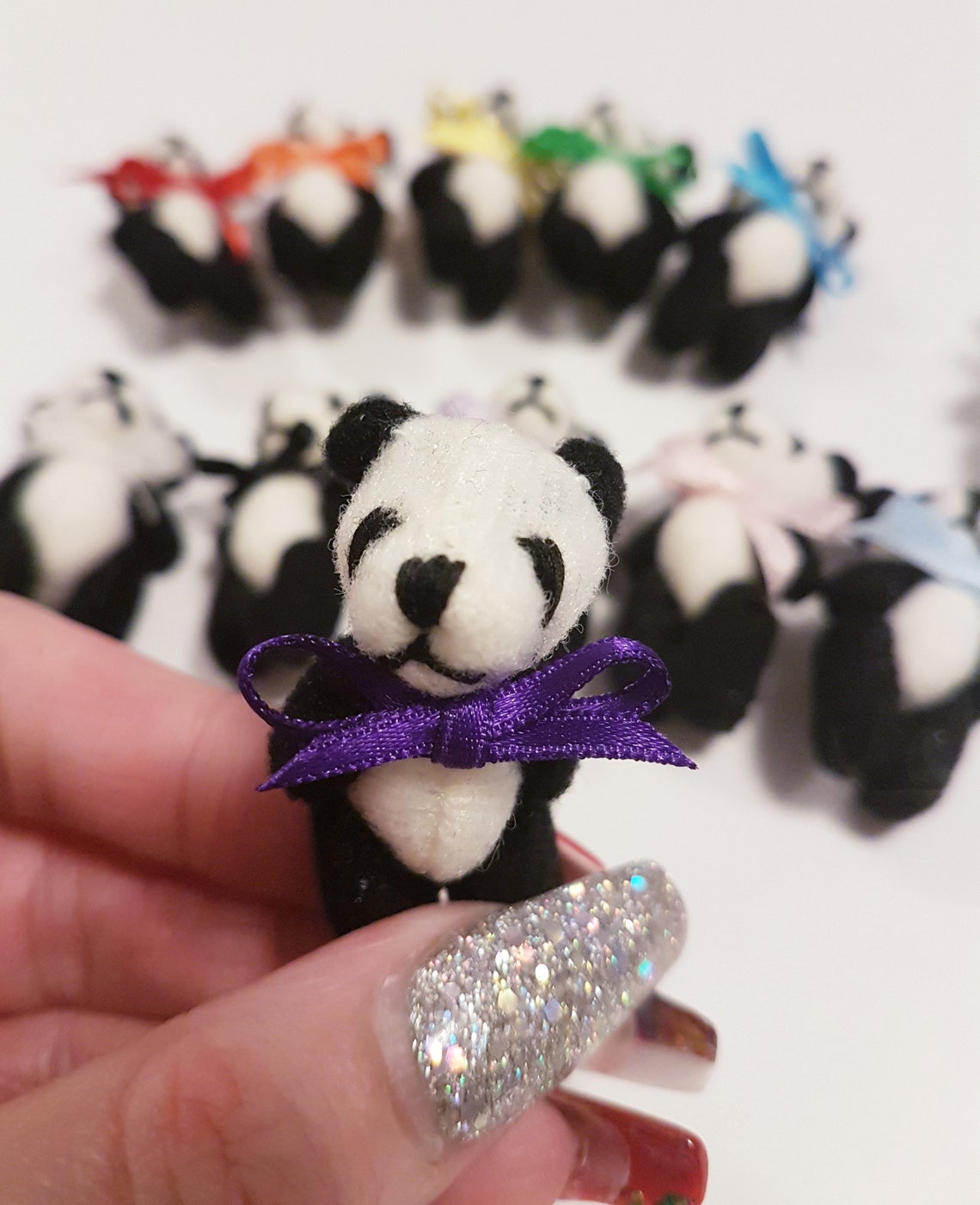 Single miniature panda teddy bear with a purple ribbon bow tie held between finger and thumb. Other panda bears with different colour bow ties can be seen in the background on white