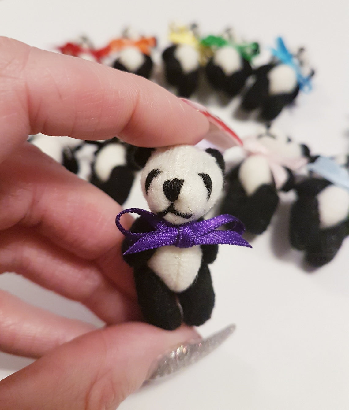 Single miniature panda teddy bear with a purple ribbon bow tie held between finger and thumb. Other panda bears with different colour bow ties can be seen in the background on white