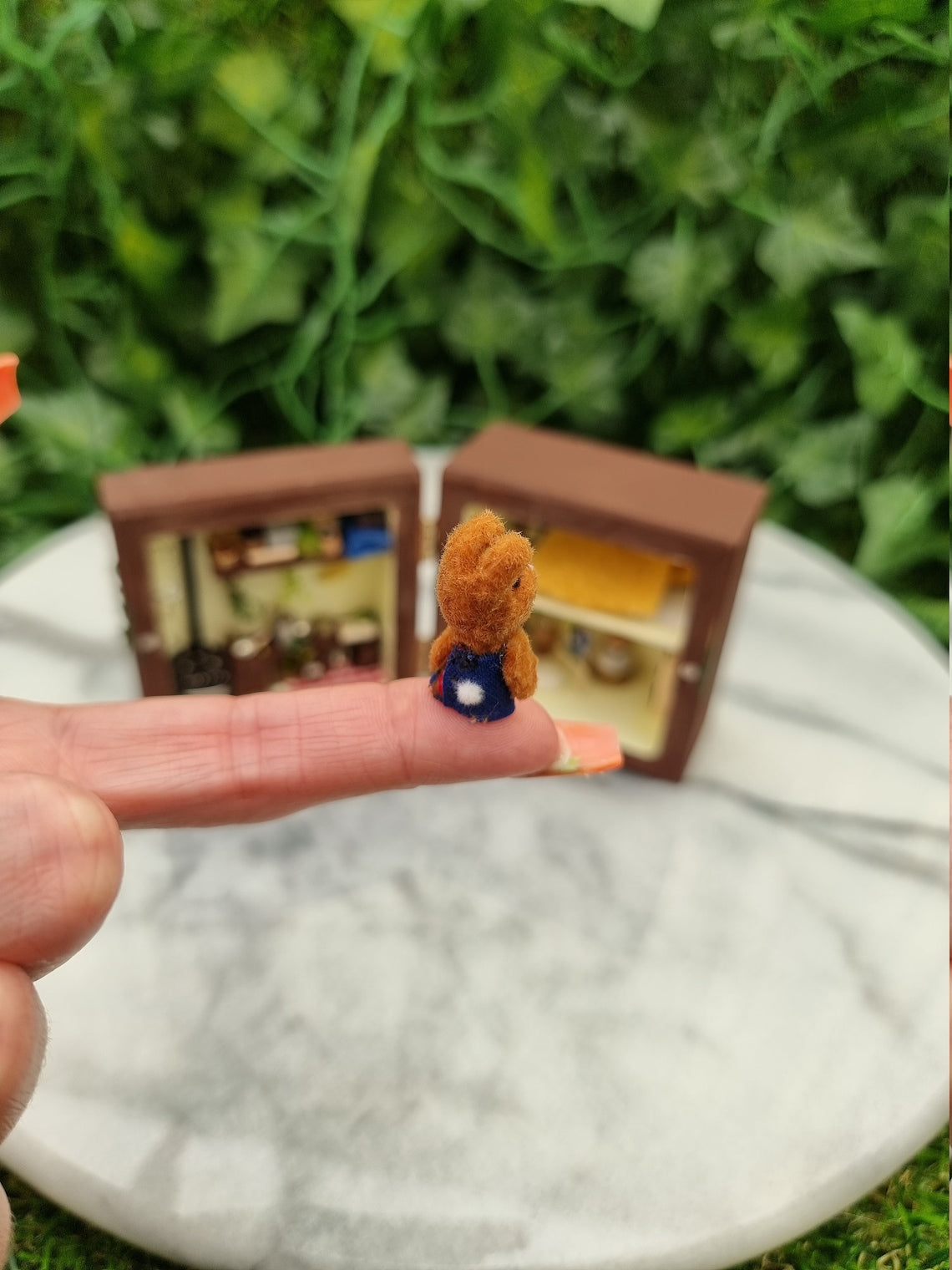 A small brown bunny in a dress sits on a finger, with a tiny cotton tail at the back of the dress.  It is held in front of a small brown box diorama open like a book in the background with a kitchen, lounge and bedroom