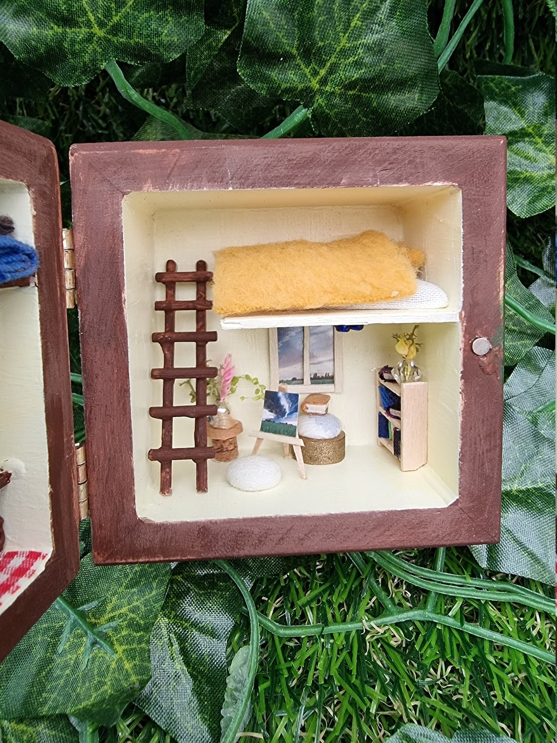 Details of a small brown box diorama. Image shows the  right side of the box diorama showing a bookcase, vases, table, easel, chairs and ladder to a loft bed with a pillow and duvet. A window looks out onto fields and clouds.