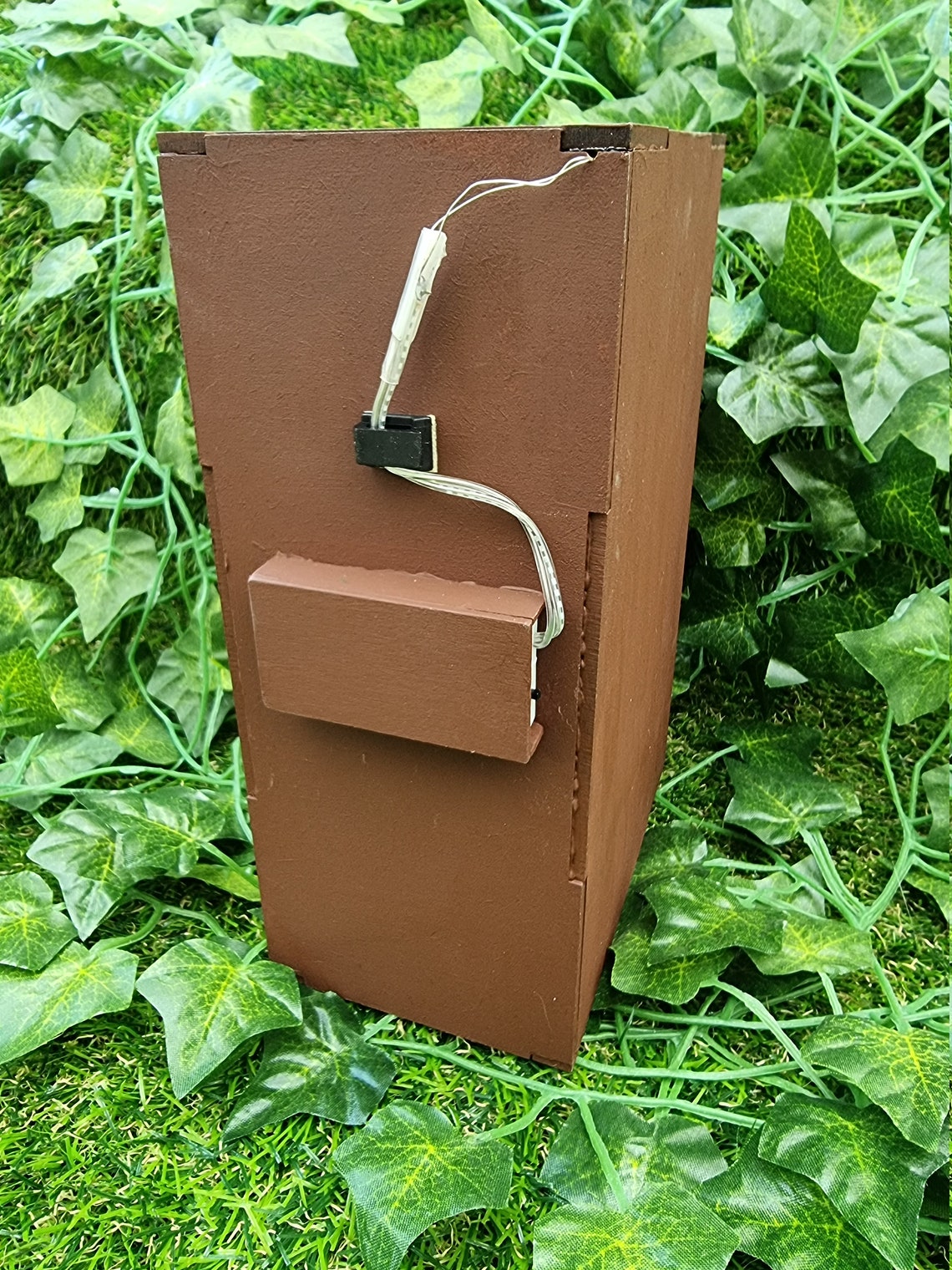 The back of the book nook box is shown. It is brown with a small insert halfway up the book nook box for a battery pack with the wire coming from the box lid. The booknook is shown on a green ivy background