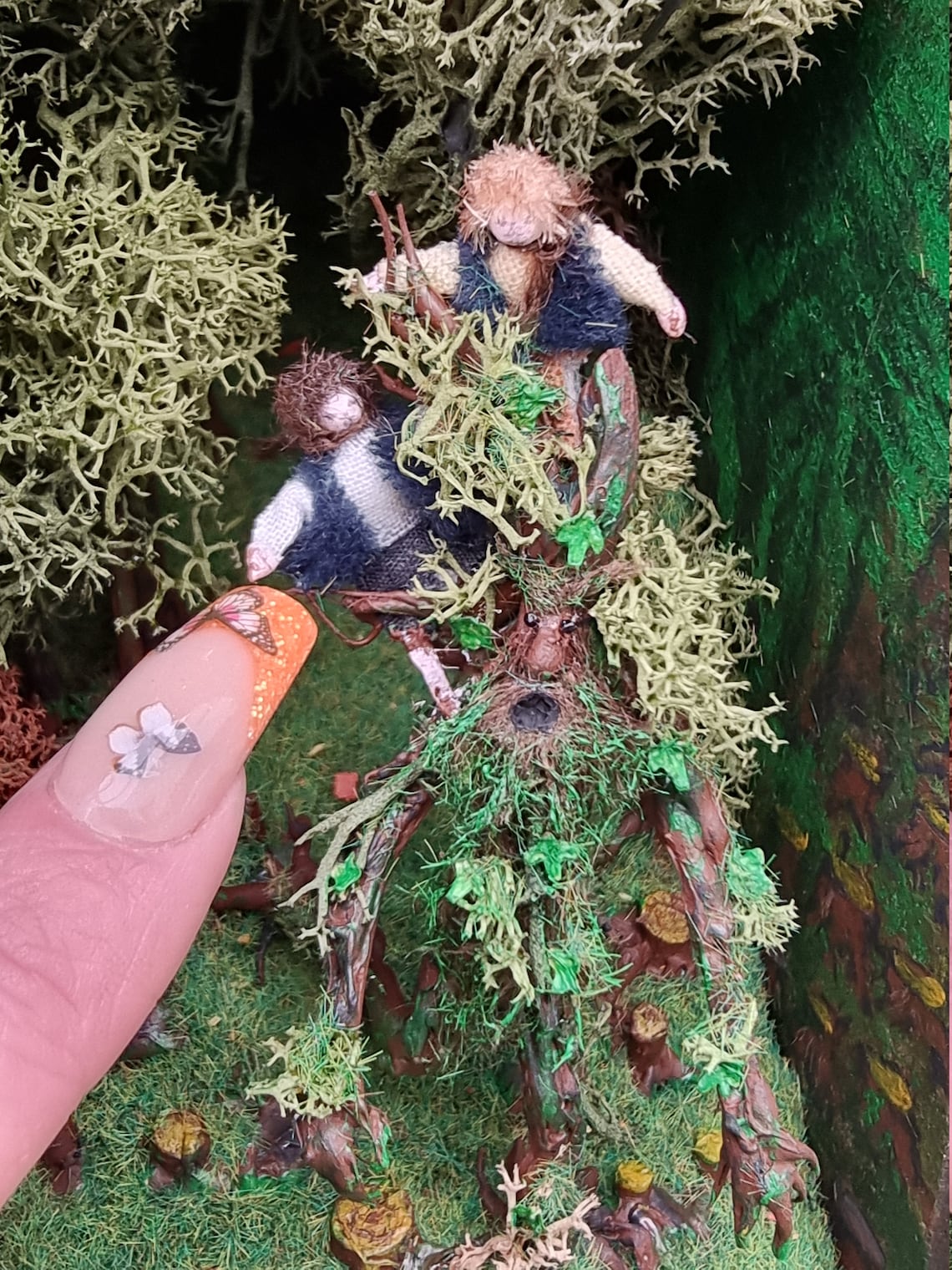 Finger points to the miniature hobbits sat on a living tree, Treebeard in the booknook scene. The hobbits have green waistcoats with white, black and brown clothing. Treebeard has a green foliage beard and other organic attachment.