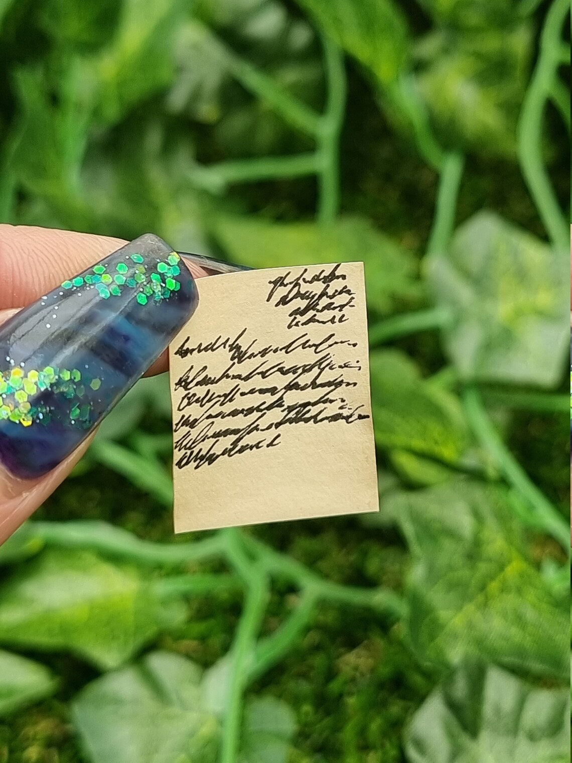 Single aged 1: 12 scale miniature letter held by a thumbnail and finger. The letters are brown, showing black text. The letter is half written. Green ivy is seen in the background.