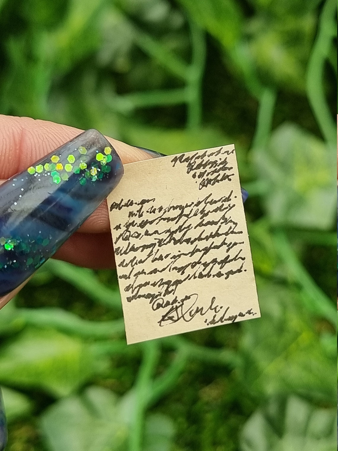 Single aged 1:12 scale miniature letters held with a thumbnail and forefinger. The letter brown with black text and is fully written. Green ivy is seen in the background