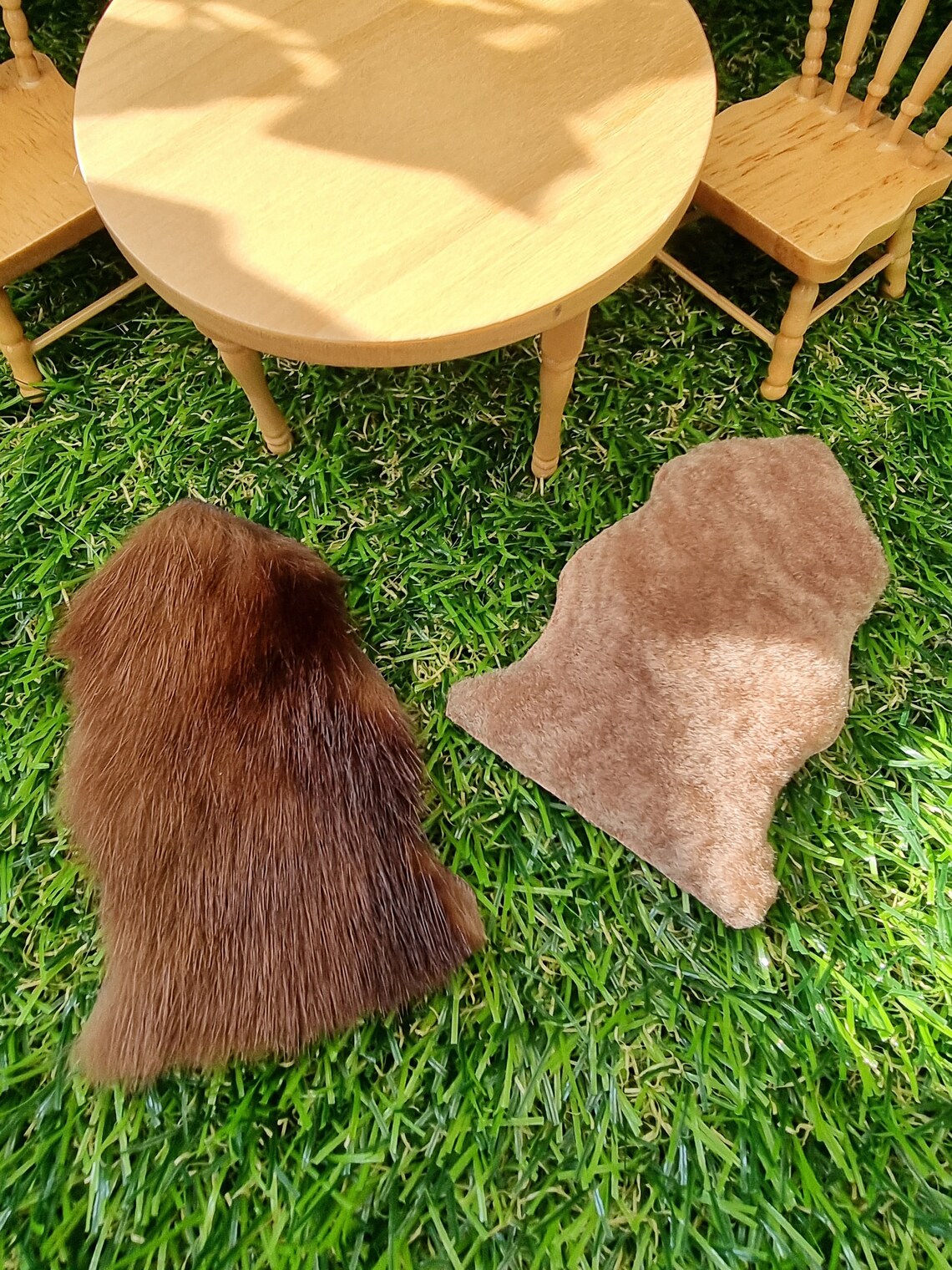 Two 1:12 scale animal skin genuine fur rugs lie on a green grass floor with miniature table and chairs in the background. One is dark brown and the other is light brown.