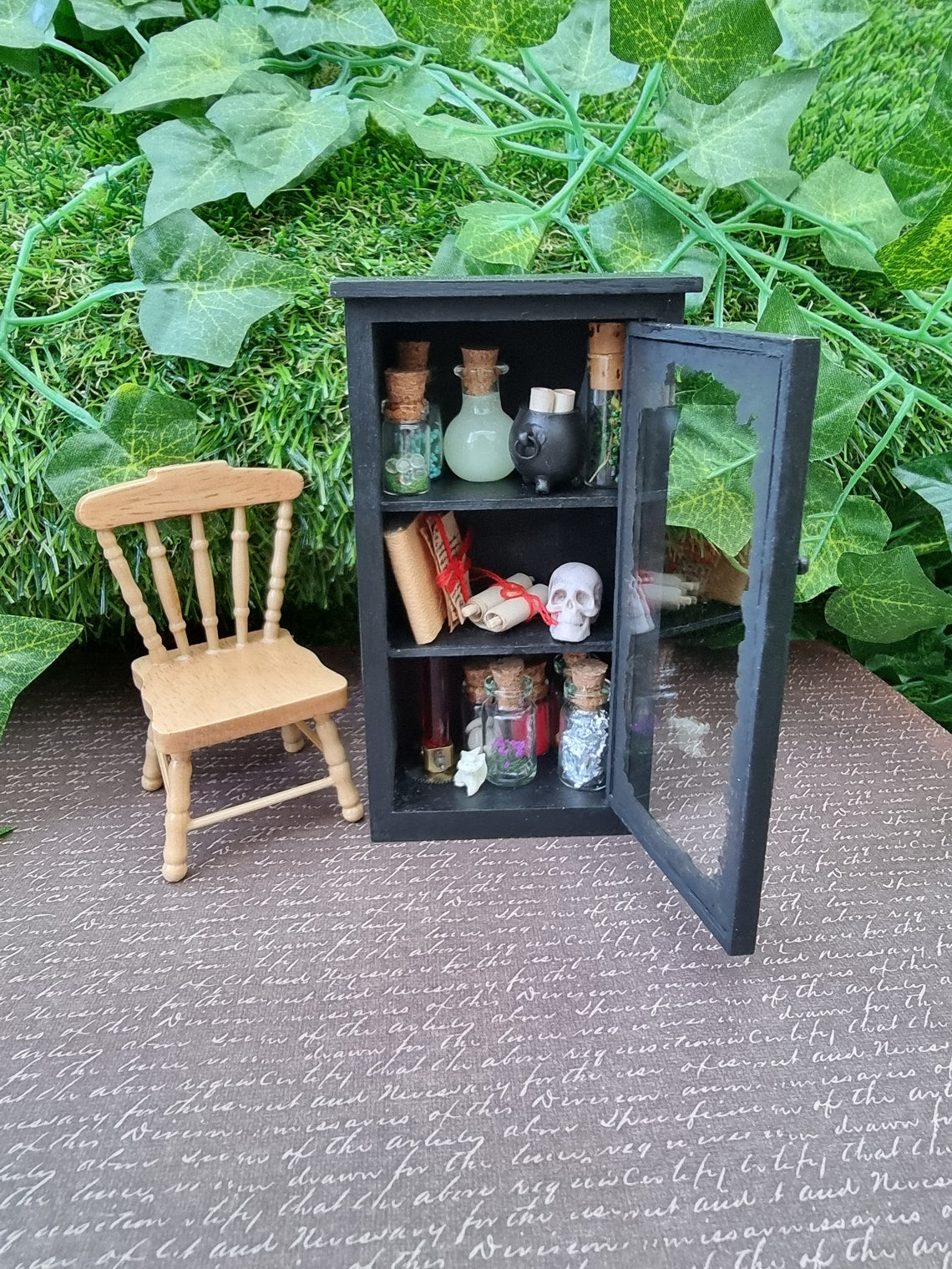 Open mini glass-fronted black apothecary cabinet stands beside a minature chair against an ivy background. The cabinet has 3 shelves. The top shelf has apothecary bottles and a cauldron. Middle shelf has a skull and scroll. Bottom shelf has bottles