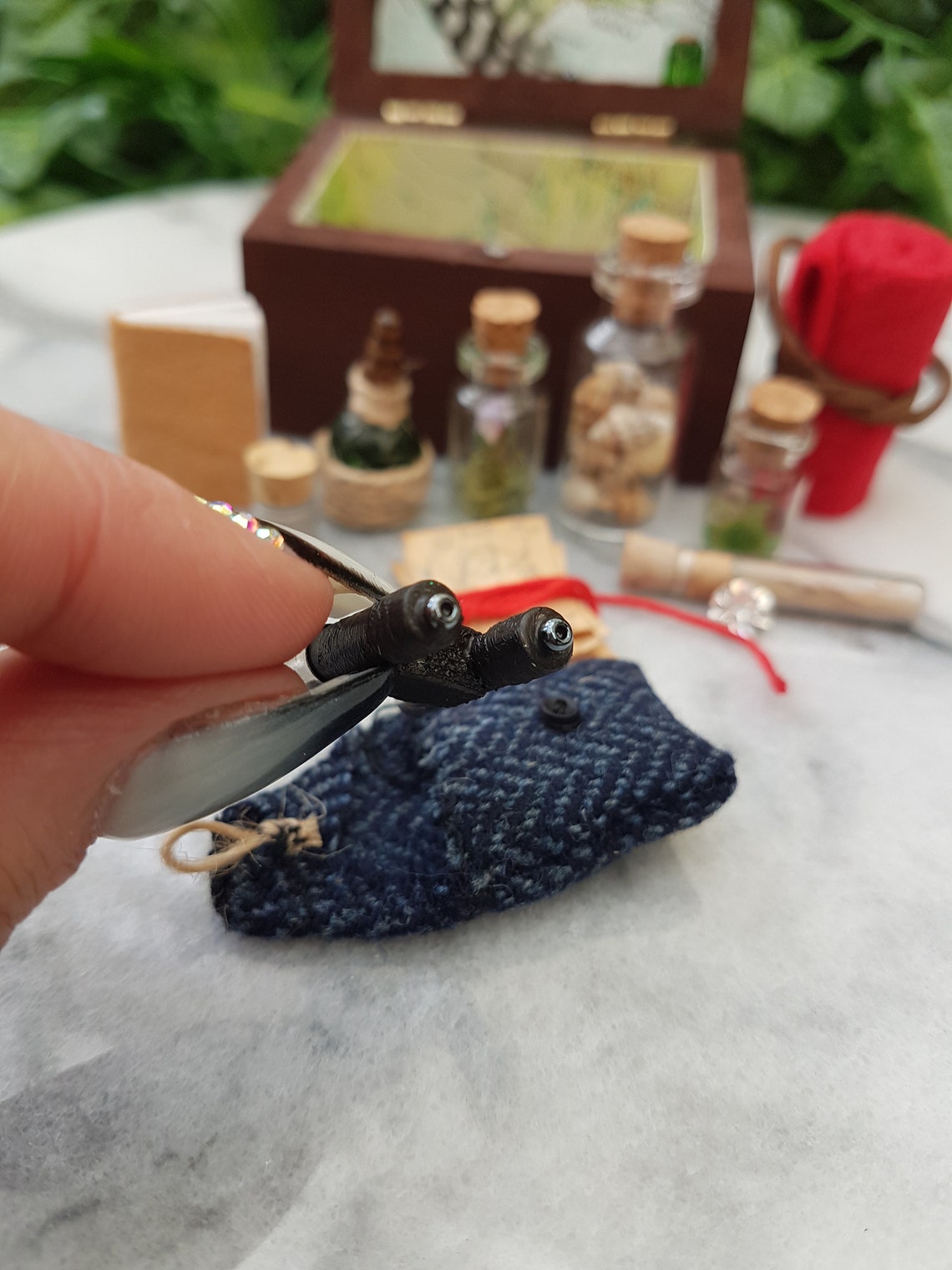 Hand holds a pair of tiny black binoculars along with a tiny tweed pouch. An open travellers chest sitsin the background glass vials, leather book, quill and inkwell, papers and red blanket roll. The inside of the chest is lined with map paper.