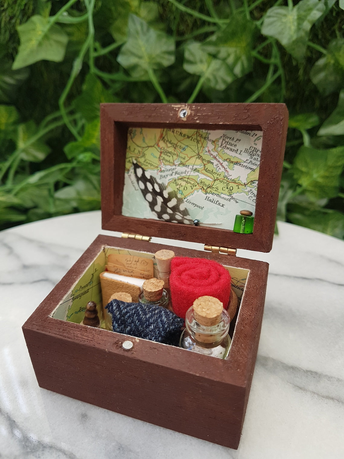 An open travellers chest is shown filled neatly with glass vials, tweed pouch, leather book, quill and inkwell, papers and red blanket roll. The inside of the chest is lined with coloured map paper. Shown on a marble slab