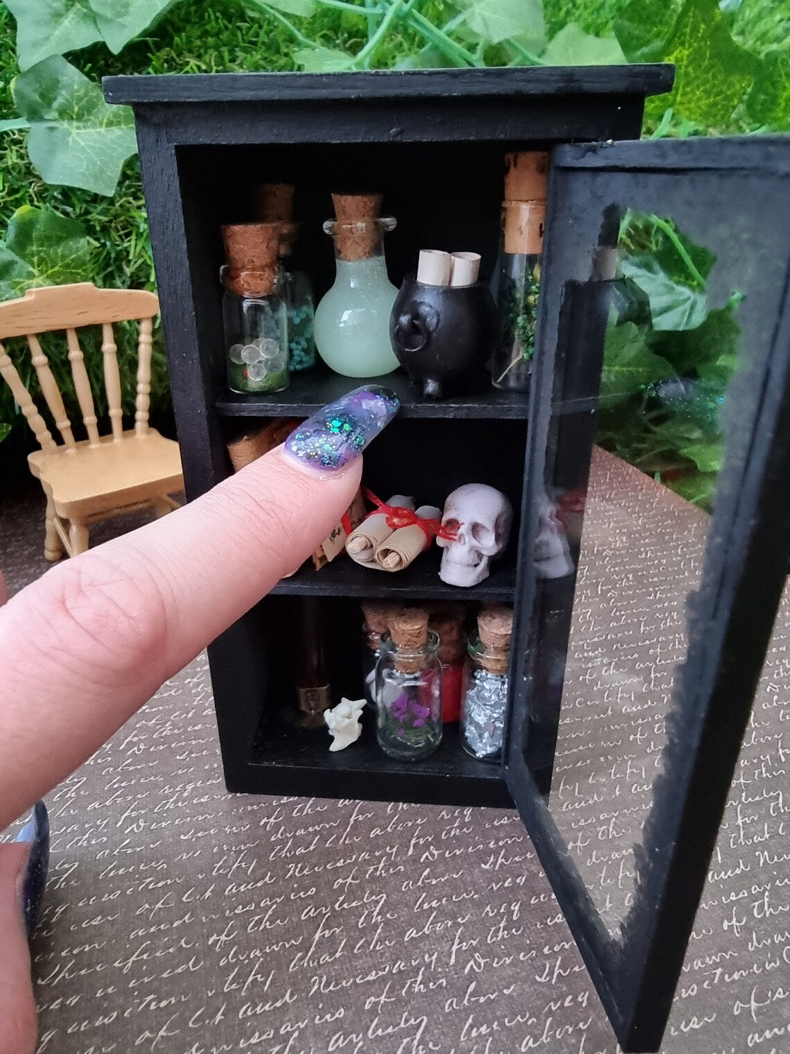 A miniature 1:12 scale glass-fronted black apothecary cabinet stands against an ivy background. The cabinet has 3 shelves. The top shelf has apothecary bottles and cauldron. Middle shelf has a skull and scroll. Bottom shelf has bottles. Finger points for scale