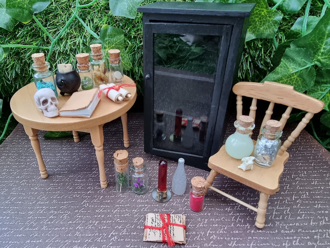 Open miniature 1:12 scale glass-fronted black apothecary cabinet stands against an ivy background. The cabinet has 3 shelves and is open and empty with contents sat beside, including apothecary bottles, a cauldron, book, skull and scroll.