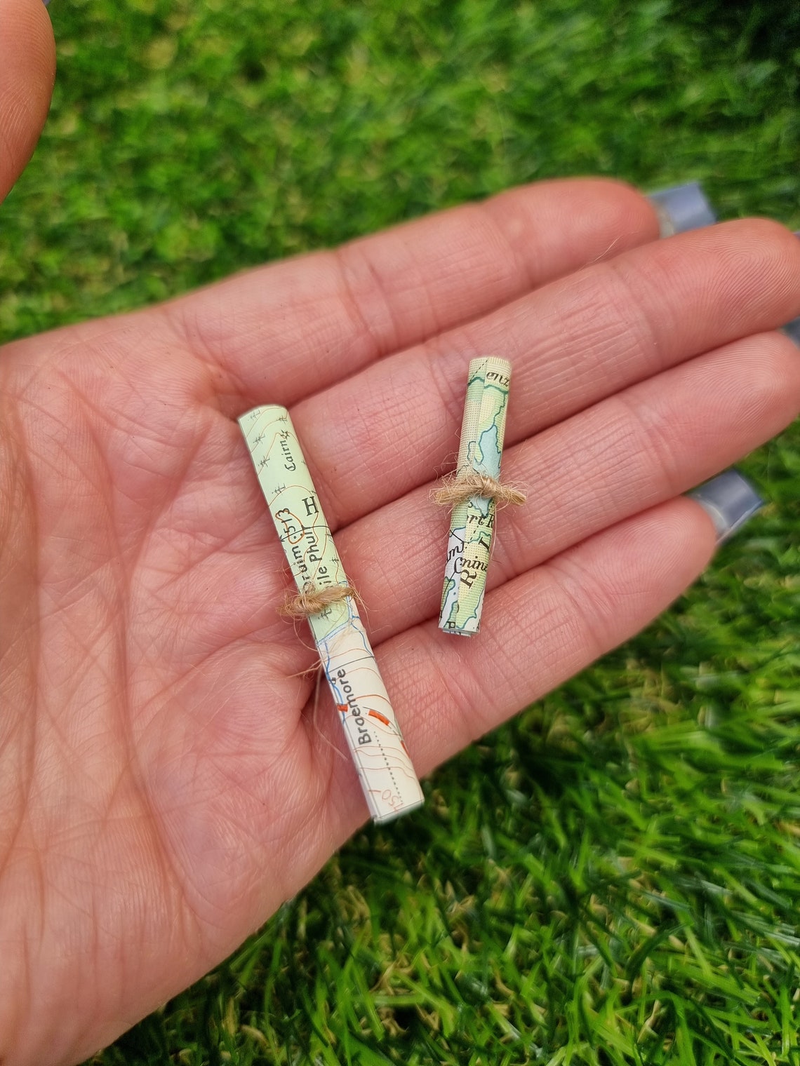 Two 1:12 scale miniature map rolls tied with jute string are displayed in the palm of a hand. One map roll is longer than the other. The maps are mainly coloured white, green and blue