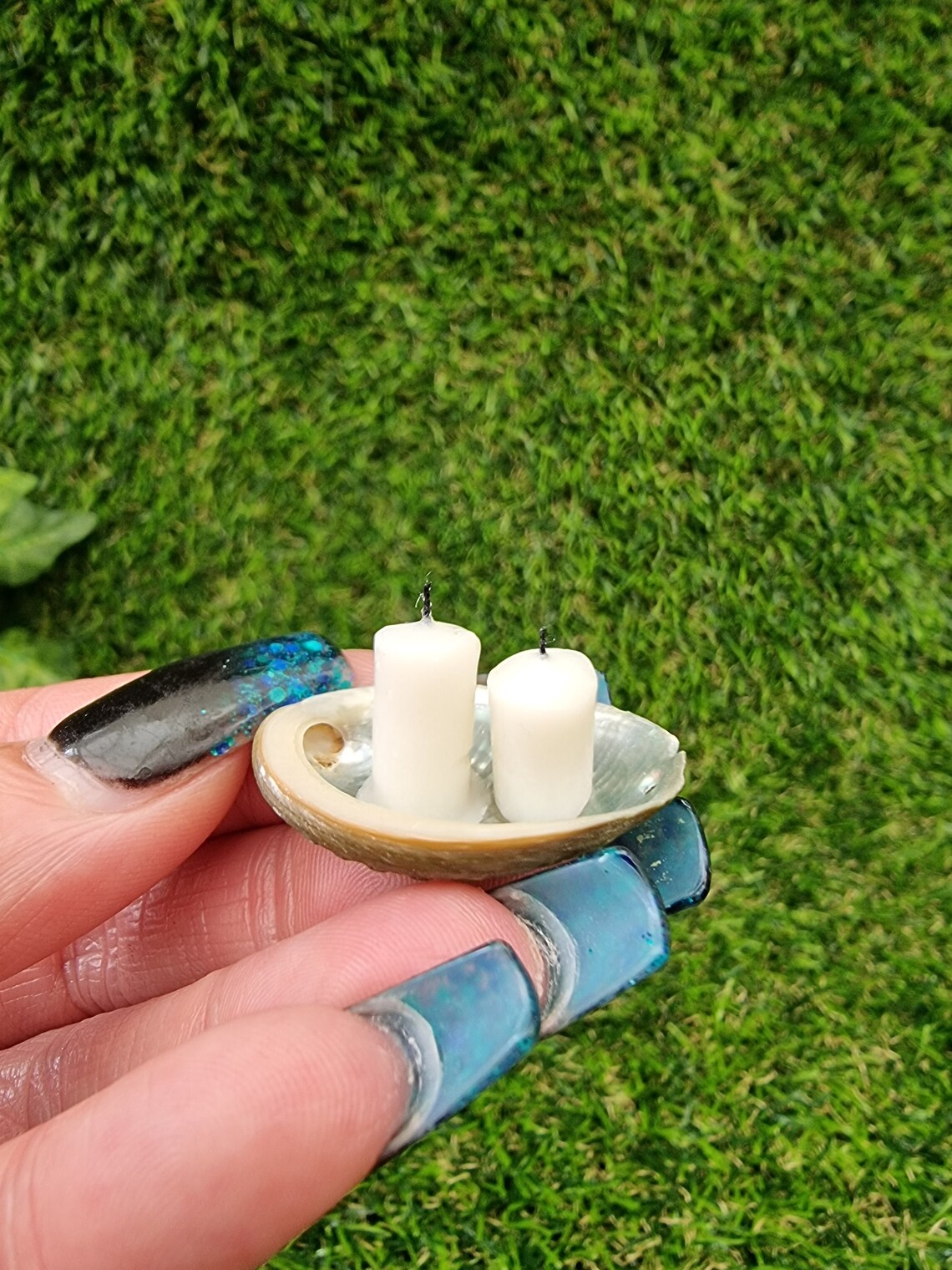 Two white 1:12 scale miniature candles in a small abalone shell dish, displayed on fingers of an open hand