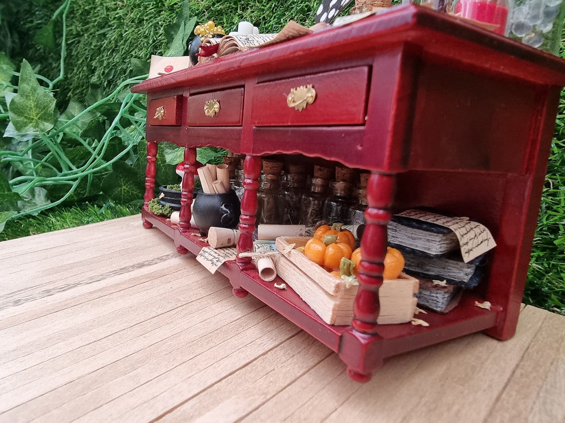 The bottom of the mahogany brown 1:12 scale miniature sideboard shows a stack of books with parchment rolls, maps rolls and a wooden crate of small orange pumpkins. Filled glass vials and a black cauldron filled with parchment rolls sits on the shelf