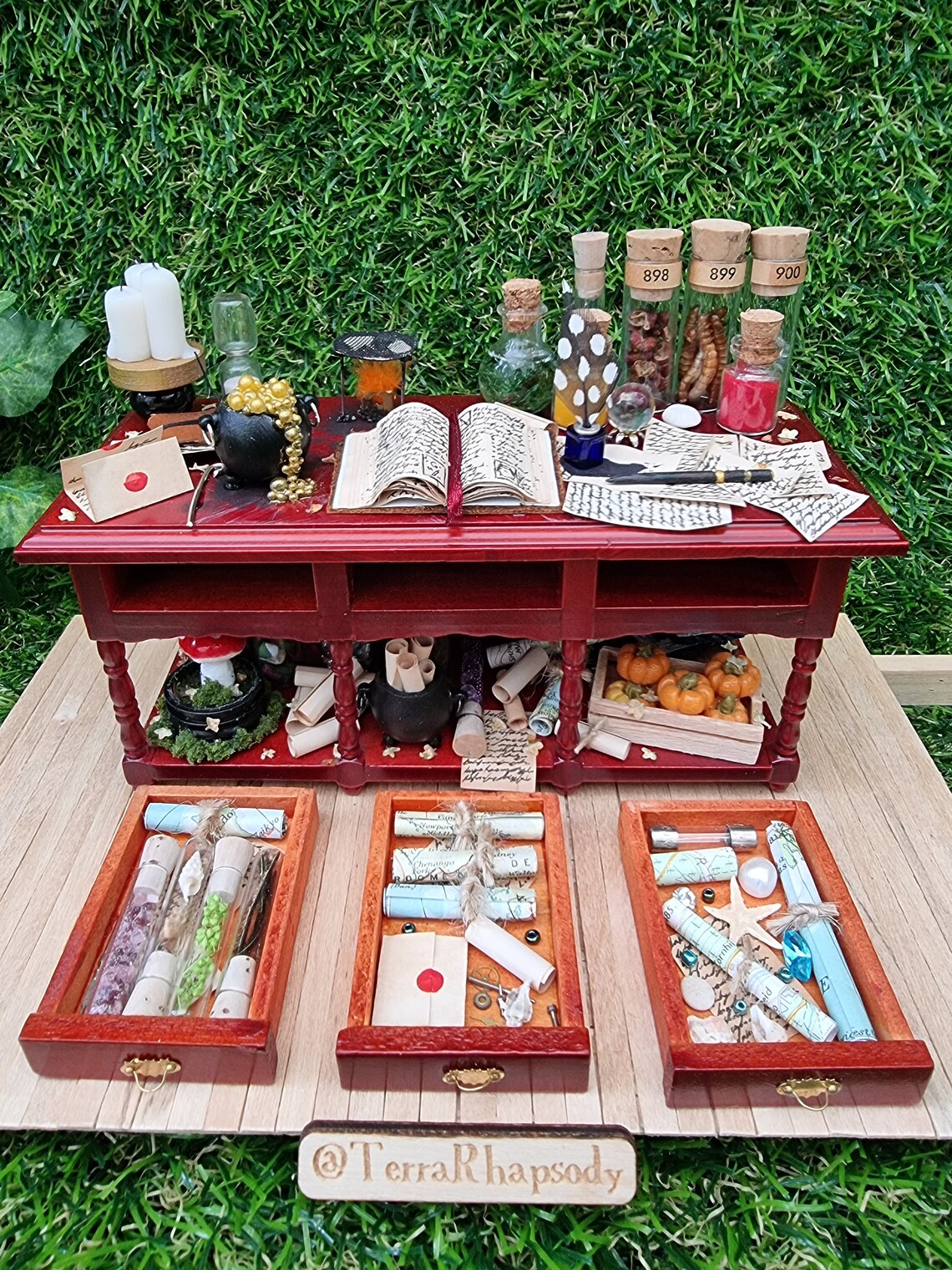 A 1:12 scale miniature brown wooden sideboard has 3 open drawers. The bench top shows a bubbling cauldron, open spellbook, filled glass vials, parchments, wand, candles, burner, sand timer, and the drawers contain glass vials, letters, maps rolls and starfish