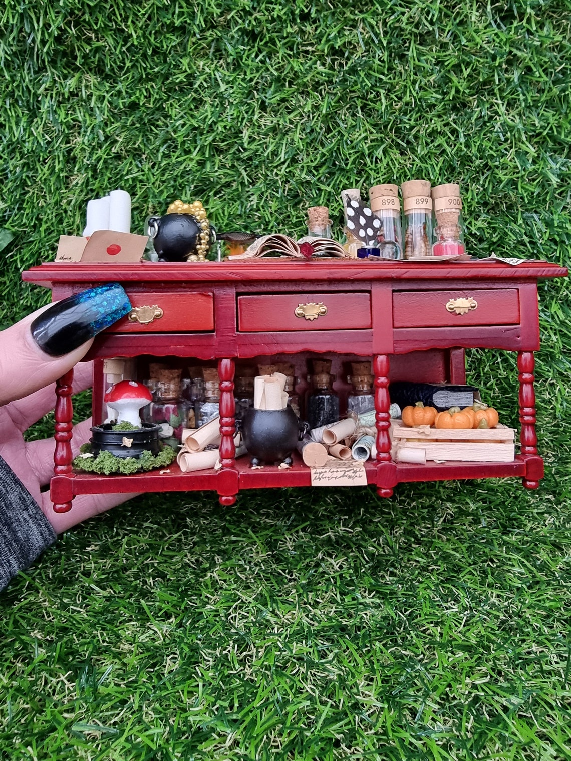 Hand holds 1:12 scale miniature brown wooden sideboard with 3 drawers and an open display area beneath. The bench hasa bubbling cauldron, open spellbook, filled glass vials, parchments, candles, map rolls, mushroom garden and a crate of pumpkins