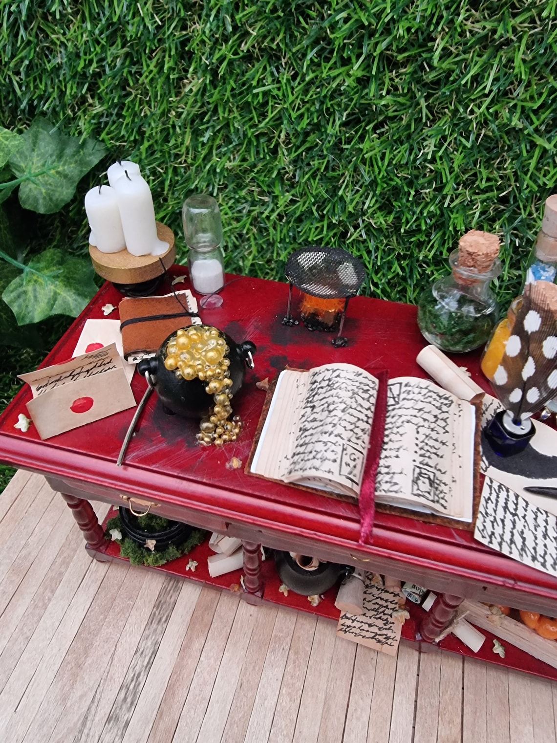 A miniature brown wooden sideboard has 3 drawers and an open display area beneath. The bench top shows a bubbling cauldron, white candles, open spellbook, leatherbound journal, filled glass vials, sand timer,  burner, letter, quill and inkwell