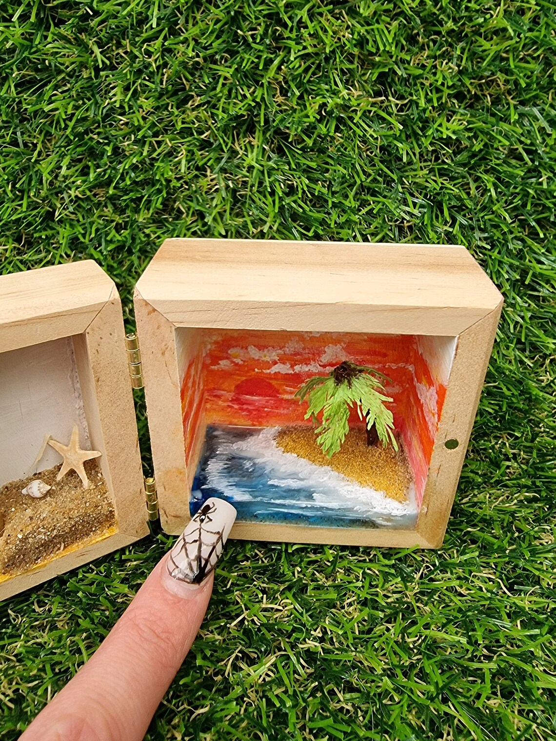 Detail of the right side of a box diorama, showing a miniature desert with a palm tree island surrounded by blue resin sea and a deep orange sunset in the background. A finger points for scale