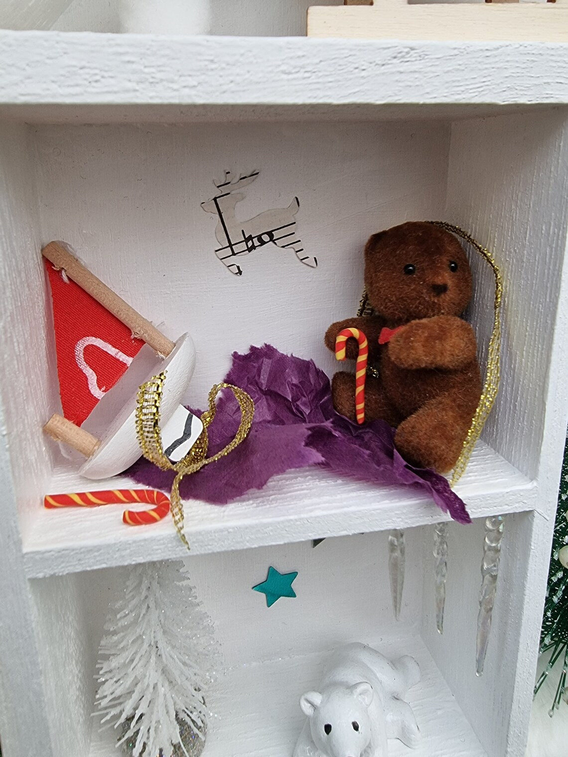 Detail of a small white shelving unit in the shape of a house. Each compartment has a different Christmas scene including a brown bear and model boat surrounded by candy canee, wrapping paper and ribbon. A polar bear and snowy tree are seen beneath