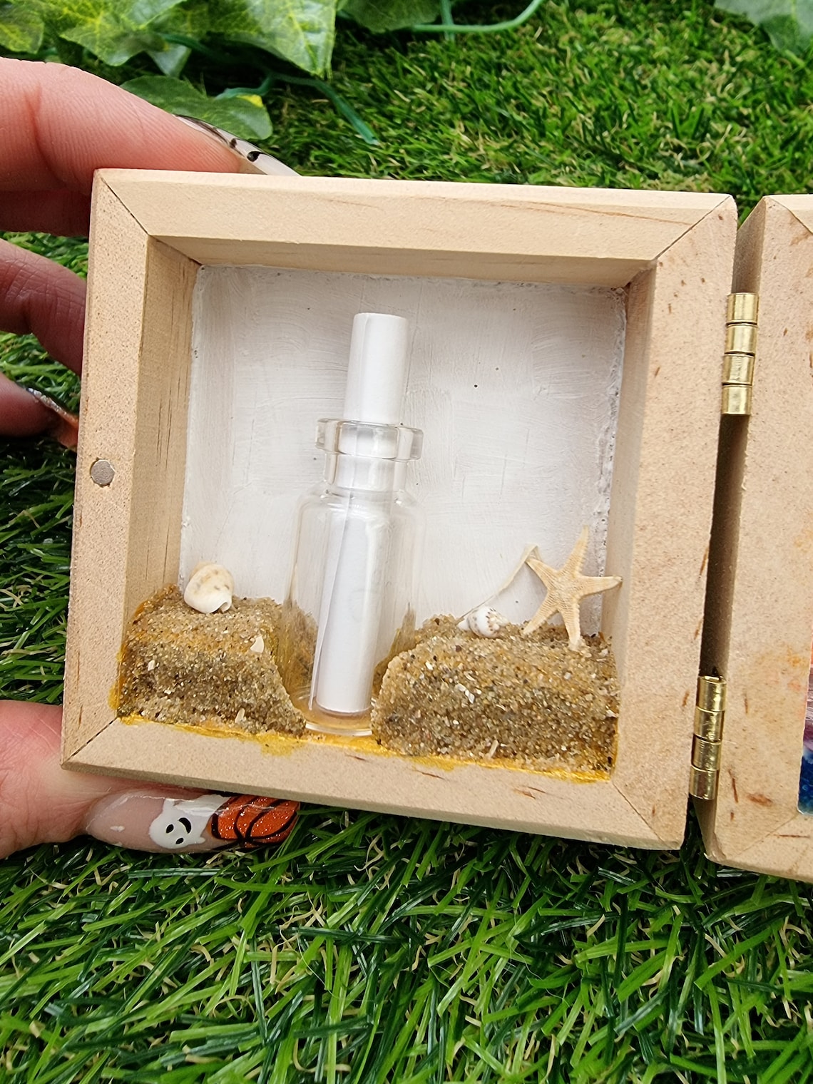 Detail of the left side of a box diorama, showing a miniature beach with an indent for a mini glass vial containing a white scroll of paper. The beach has shells and a starfish. A hand holds the box