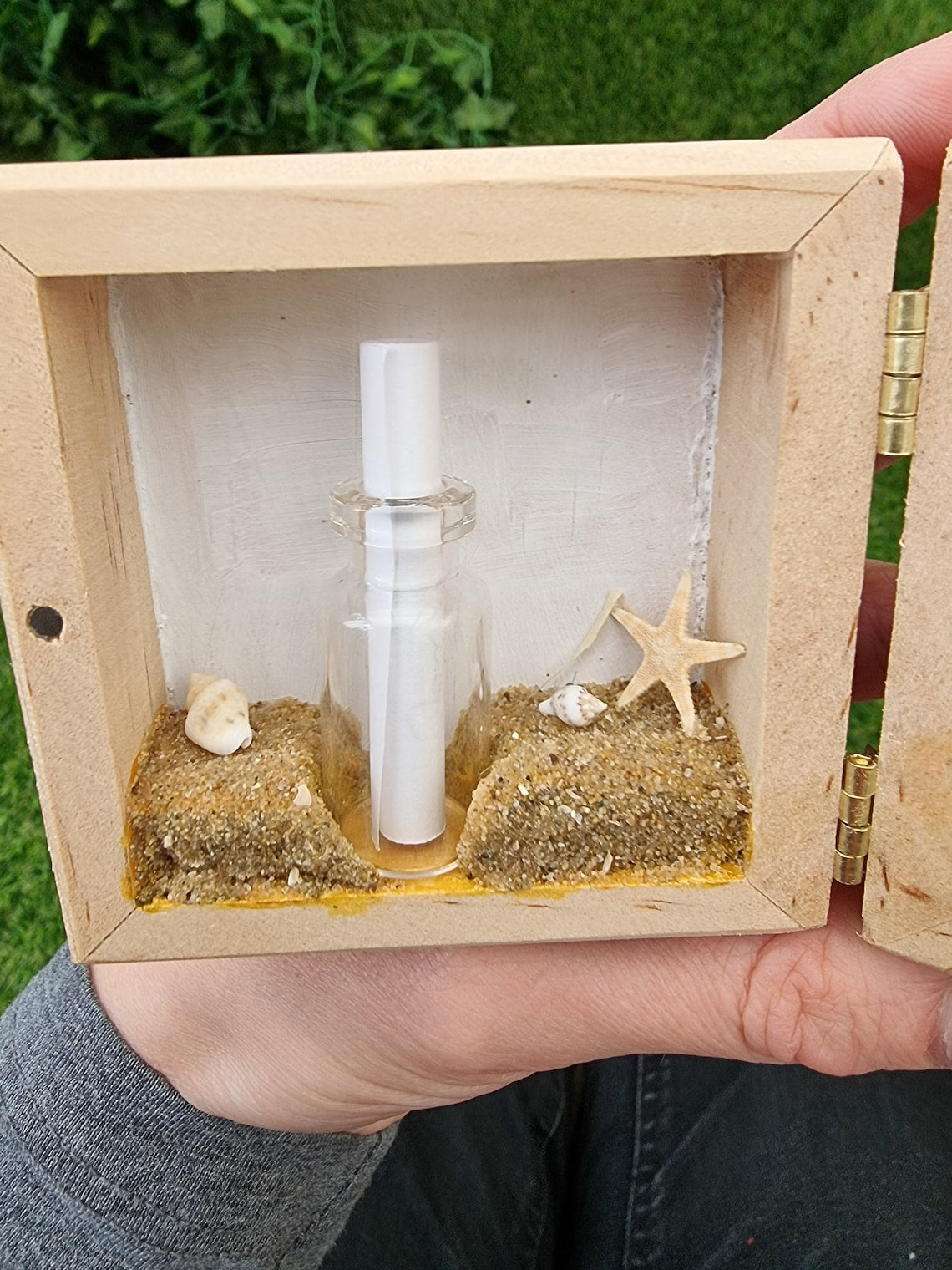 Detail of the left side of a box diorama, showing a miniature beach with an indent for a mini glass vial containing a white scroll of paper. The beach has shells and a starfish. A hand holds the box