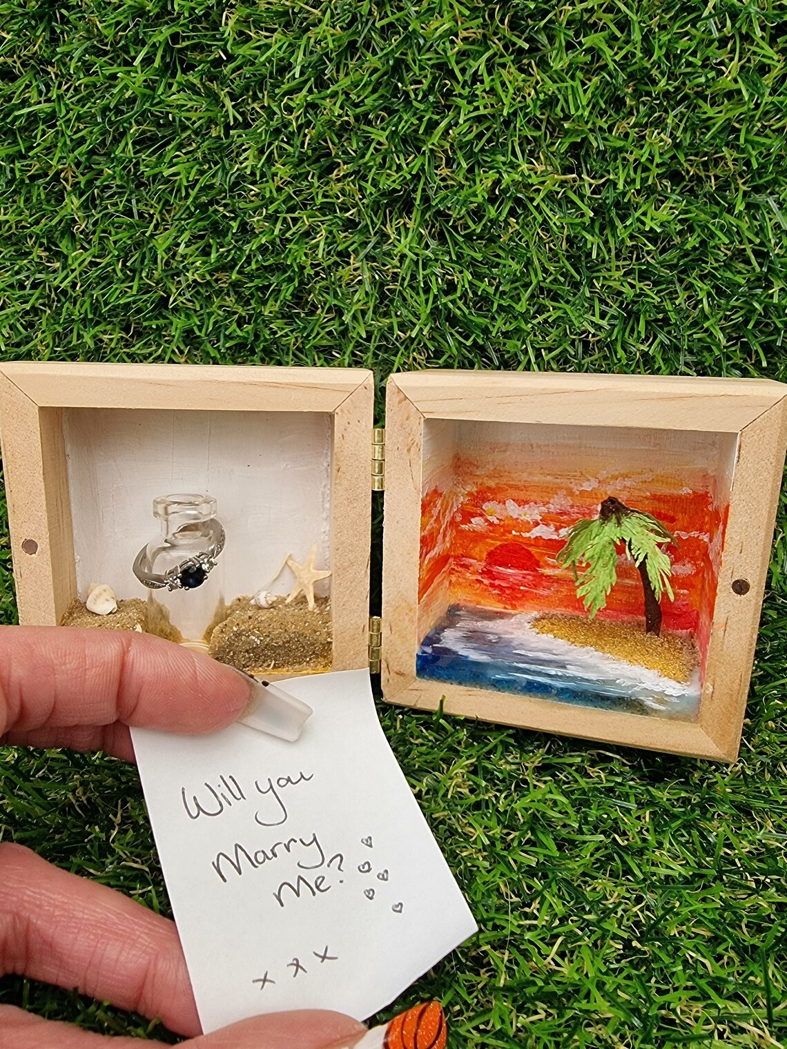 Detail of the left side of a box diorama, showing a miniature beach with an indent for a mini glass vial containing a white scroll of paper. The beach has shells and a starfish. A hand holds the box