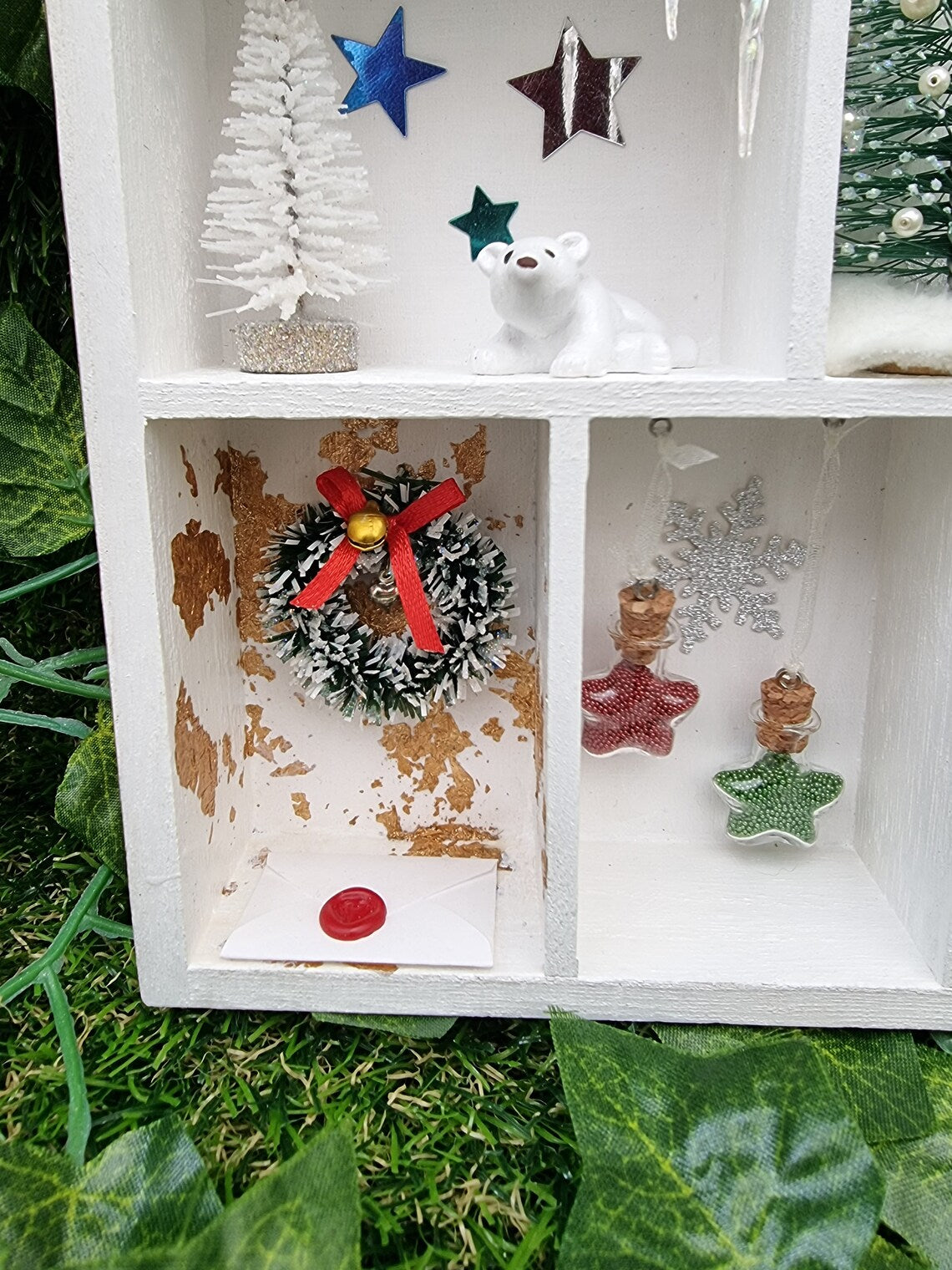 Detail of a small white shelving unit in the shape of a house. Each compartment has a different Christmas scene including green wreath with red bow and gold bell and wax sealed letter and hanging glass star baubles in red and green