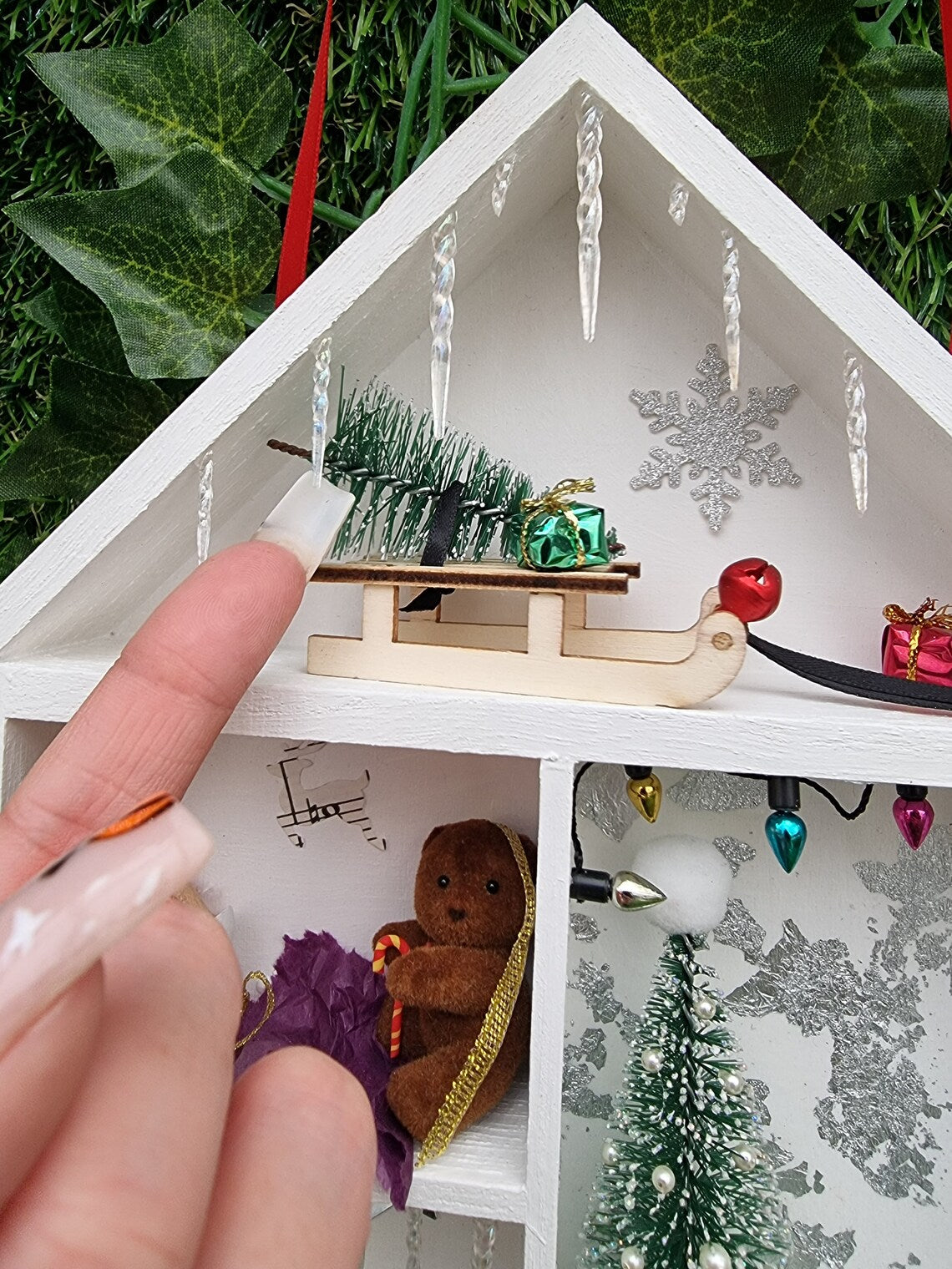 Detail of a small white shelving unit in the shape of a house. Each compartment has a different Christmas scene. A finger points to a small sledge with a christmas tree. Icicles line the roof. A christmas tree and gifts are seen beneath