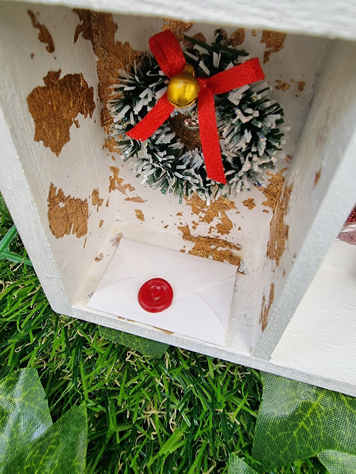 Detail of a small white shelving unit in the shape of a house. Each compartment has a different Christmas scene including green wreath with a red bow and gold bell, gold leaf background and wax sealed letter